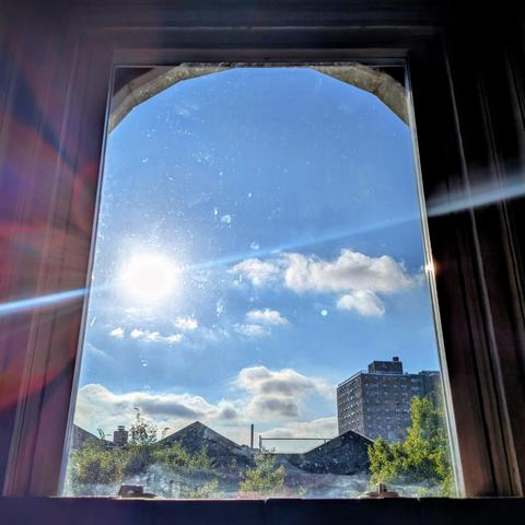 Looking through an arched window two hours after sunrise the sun is above the buildings on the left, shining directly into the lens, creating a complex lens flare. The blue sky is full of fluffy white clouds. Pointed roofs of Harlem brownstones are silhouetted across the street, and a taller apartment building can be seen in the distance. The green tops of two trees are on the bottom and right.