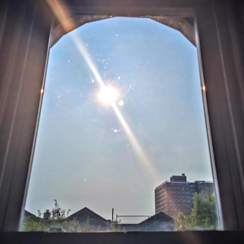 Looking through an arched window two-and-a-half hours after sunrise the sun is just left of center in the window, shining directly into the lens, creating a diagonal lens flare and illuminating the dirt and scratches on the glass.  The sky is robin egg blue and cloudless. Pointed roofs of Harlem brownstones are silhouetted across the street, and a taller apartment building can be seen in the distance. The green tops of two trees are on the bottom and right.