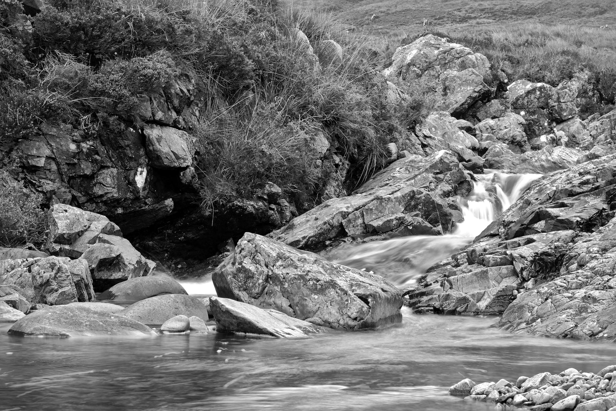 A black-and-white photograph, high contrast. In the foreground, there is moving water (long exposure, the movement in the surface can be seen). There are some small rocks in the front right and larger rocks throughout the width of the picture where the foreground body of water meets the upper half of the picture. Above on the right, a small stream comes down over rocks (flowing faster than the water surface in the foreground). Above the rocks leading the water toward the foreground, there are hilly grasslands in the distance. On the left side, the upper half of the image is made up of a rock formation with grass growing on top. A black-and-white photograph, high contrast. In the foreground, there is moving water (long exposure, the movement in the surface can be seen). There are some small rocks in the front right and larger rocks throughout the width of the picture where the foreground body of water meets the upper half of the picture. Above on the right, a small stream comes down over rocks (flowing faster than the water surface in the foreground). Above the rocks leading the water toward the foreground, there are hilly grasslands in the distance. On the left side, the upper half of the image is made up of a rock formation with grass growing on top.