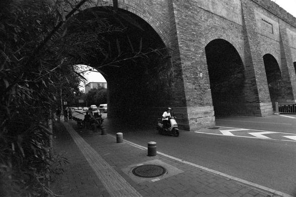 Lucky Lucky SHD 400 (FF)

English Alt Text:
A black-and-white urban street scene featuring a large brick archway structure, possibly a bridge or viaduct, with multiple arches. A person rides a scooter toward the camera on a curving road. On the left, pedestrians walk along a sidewalk lined with bollards and foliage. The moody lighting and nostalgic tone evoke a sense of quiet city life.

中文替代文字：
这是一张黑白街景照片，右侧是一座大型砖砌拱门结构，可能是桥梁或高架桥，有多个拱洞。一人骑着踏板车沿弯曲道路向镜头驶来。左侧人行道上有行人，路边设有防撞柱并被绿植遮掩。画面光线昏暗，氛围怀旧，展现了城市…