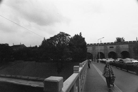 Lucky Lucky SHD 400 (FF)

English Alt Text:
A black-and-white photo of a pedestrian bridge leading to a historic stone structure with multiple archways and battlements. The bridge has railings on both sides and a few people walking across. Parked cars and a streetlamp line the right side of the scene. Trees surround the stone structure, and the sky is overcast, adding a moody atmosphere. The contrast between the ancient architecture and modern elements like vehicles and lighting evokes a sense …