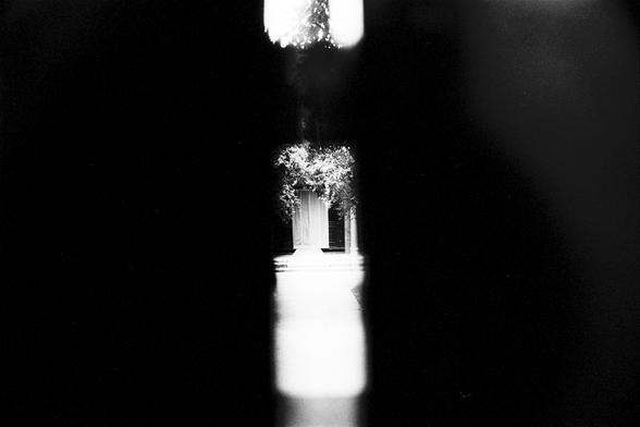 Lucky Lucky SHD 400 (FF)

English Alt Text:
A black-and-white photograph taken through a narrow vertical slit, resembling a keyhole. The foreground is completely dark, framing a bright, well-lit doorway in the center. The doorway is surrounded by hanging vines or foliage, suggesting a garden or secluded courtyard. The contrast between the dark framing and the illuminated scene creates a sense of mystery, as if the viewer is peeking into a hidden or private space. The composition draws the eye d…