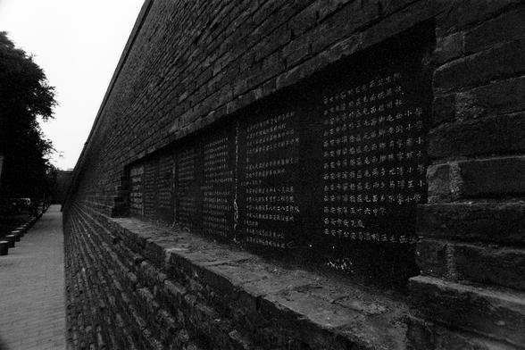 Lucky Lucky SHD 400 (FF)

English Alt Text:
A black-and-white photograph of a long brick wall embedded with multiple black stone plaques. Each plaque is inscribed with vertical columns of Chinese characters, possibly historical or commemorative text. The wall stretches into the distance, creating depth and perspective. A paved walkway runs alongside the wall, and trees are visible in the background. The monochrome tone adds a solemn and reflective atmosphere, suggesting the site may be of cultu…