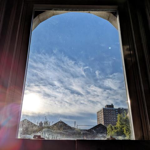 Looking through an arched window an hour after sunrise the blue sky is mostly filled with  swirling white clouds. The sun is entering on the left, creating a lens flare. Pointed roofs of Harlem brownstones are silhouetted across the street, and a taller apartment building can be seen in the distance. The green tops of two trees are on the bottom and right.