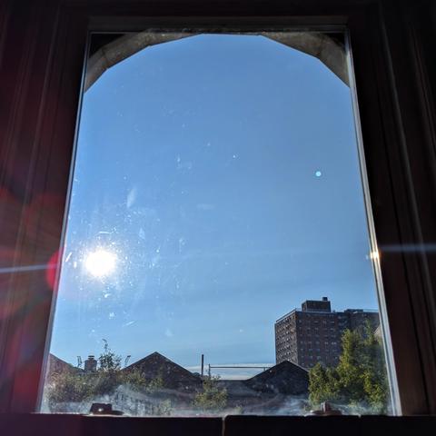 Looking through an arched window ninety minutes after sunrise the sun is above the buildings on the left in the blue sky, making a  lens flare with multi-colored petals. Pointed roofs of Harlem brownstones are silhouetted across the street, and a taller apartment building can be seen in the distance. The green tops of two trees are on the bottom and right.