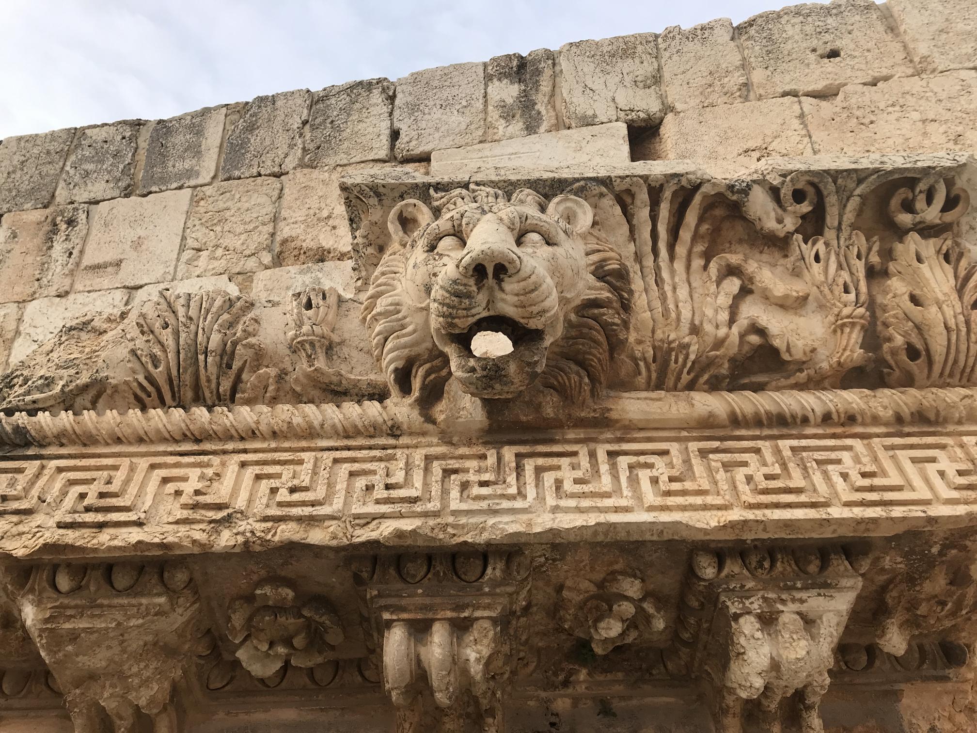 looking directly up into the mouth of a waterspout shaped like a lion's head. there are weathered floral and geometrical designs on the surrounding structure.
