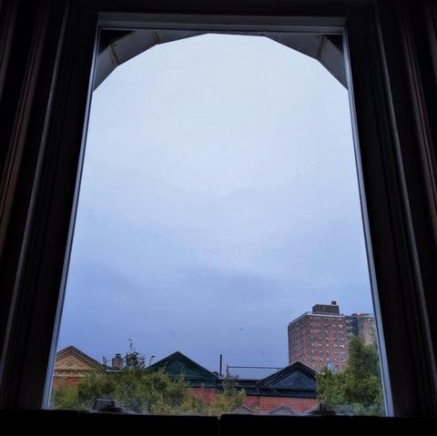 Looking through an arched window an hour after sunrise the sky is full of grey rain clouds. Pointed roofs of Harlem brownstones with red brickwork are across the street, and a taller apartment building can be seen in the distance. The green tops of two trees are on the bottom and right.