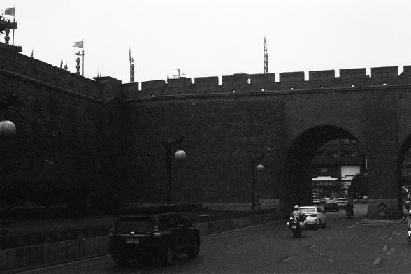 Lucky Lucky SHD 400 (FF)

English Alt Text:
A black-and-white image captures a busy street passing through a large stone wall with battlements and flags. Two arched gateways allow cars and motorcycles to pass. The scene blends ancient architecture with modern urban life.

中文替代文字：
这张黑白照片展示了一条繁忙街道穿过一座带有城垛和旗帜的巨大石墙。墙上有两个拱形门洞，车辆和摩托车穿行其中。画面融合了古代建筑与现代城市生活。