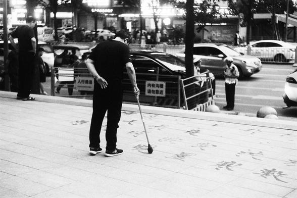Lucky Lucky SHD 400 (FF)

English Alt Text:
A black-and-white photo shows a man practicing Chinese calligraphy on a city sidewalk using a large brush dipped in water. He bends slightly forward, one hand behind his back, the other guiding the brush with precision. The wet characters shimmer briefly on the pavement before fading. Behind him, cars pass on a busy street, a police officer stands nearby, and pedestrians walk past storefronts. The scene blends fleeting traditional art with modern urba…