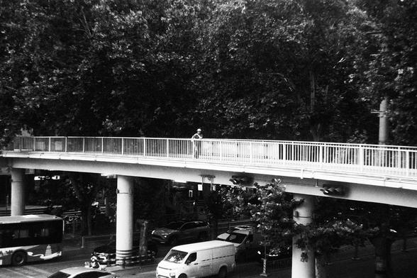 Lucky Lucky SHD 400 (FF)

English Alt Text:
A solitary figure walks across an elevated pedestrian walkway in a black-and-white cityscape. The walkway is supported by thick columns and overlooks a busy street filled with cars, buses, and a white van. Trees line the road, casting shade. The image contrasts the quiet solitude above with the bustling traffic below.

中文替代文本：
黑白城市景观中，一名行人独自走在高架人行天桥上。天桥由粗大的柱子支撑，下方是车流密集的街道，有汽车、公交车和一辆白色货车。道路两旁树木成行，投下阴影。画面呈现出天桥上的宁静与桥下喧嚣交通之间的对比。
