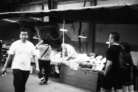 Lucky Lucky SHD 400 (FF)

English Alt Text:
In a black-and-white street market scene, a vendor leans over a table filled with produce under hanging lights. Handwritten signs in Chinese mark prices. Shoppers walk past, some browsing. A canopy shades the stall, and a tiled roof edge is visible above. The image evokes daily life and commerce, with a nostalgic tone from its monochrome palette.

中文替代文本：
黑白街市场景中，一位摊贩在挂灯下俯身整理摊位上的农产品。桌上有手写的中文标价牌。行人穿梭，有人驻足选购。摊位上方有遮阳篷，画面顶部可见瓦片屋檐。整张照片以怀旧的黑白色调呈现日常生活与市井交易的氛…