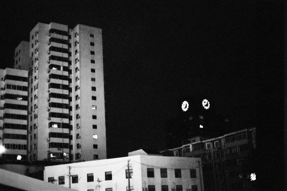 Lucky Lucky SHD 400 (FF)

English Alt Text:
A nighttime cityscape in black and white features several buildings. On the left, tall residential or office towers with lit windows rise into the dark sky. In the foreground, a low building with utility lines adds depth. On the right, a darker structure displays two glowing circular clock faces, suggesting a clock tower. The contrast between light and shadow creates a quiet, contemplative mood.

中文替代文本：
这是一张黑白城市夜景照片。左侧是几栋高层住宅或办公楼，窗户中透出灯光，直指夜空。前景中有一座低…