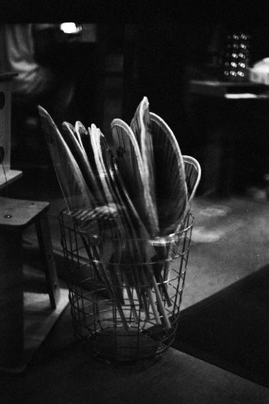Lucky Lucky SHD 400 (FF)

English Alt Text:
A black-and-white photograph shows a wire basket filled with handheld fans. The fans are arranged with their handles pointing downward and their circular heads fanning outward like petals. Each fan has a mesh or slatted surface, and they vary slightly in shape and size. The basket rests on a smooth indoor floor, possibly in a communal or recreational space. The background is softly blurred, with indistinct furniture and objects. The monochrome tones a…