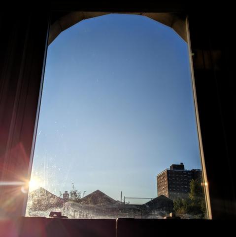 Looking through an arched window forty-five minutes after sunrise the sun is above the buildings on the left in the cloudless blue sky, making a lens flare with multi-colored petals. Pointed roofs of Harlem brownstones are silhouetted across the street, and a taller apartment building can be seen in the distance. The green tops of two trees are on the bottom and right.