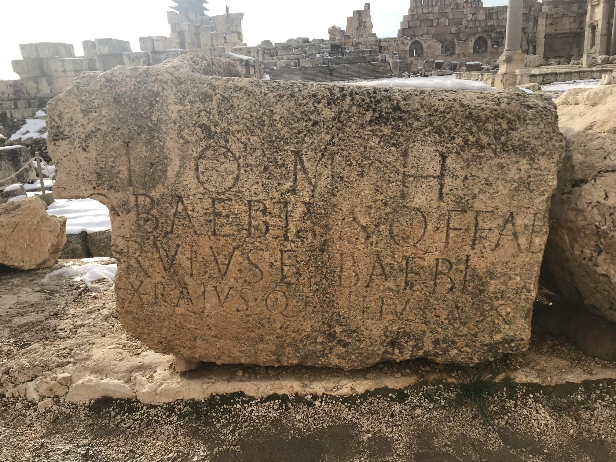 Large, weathered stone with ruins in the background. On the stone are four lines of Latin inscription in large capitals.