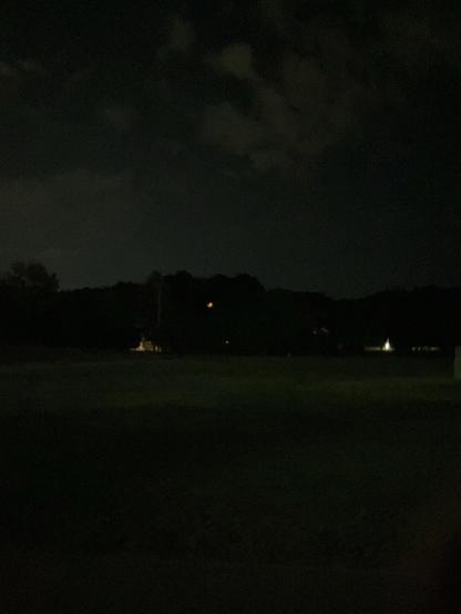A dark night landscape featuring a field with a few faint lights in the distance, surrounded by trees and a cloudy sky.