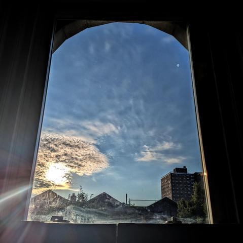 Looking through an arched window an hour after sunrise the sun is above the buildings on the left, making a lens flare that appears as a single ray of light. The sky is deep blue with a cluster of tightly textured white clouds around the sun, and gaseous textures above. Pointed roofs of Harlem brownstones are silhouetted across the street, and a taller apartment building can be seen in the distance. The window is grimy on the bottom. 