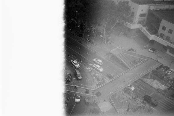 FUJIFILM NEOPAN F (FF)

English Alt Text: A black-and-white aerial photo of a city street with a unique Y-shaped pedestrian overpass crossing above the road. Cars travel in both directions below, while a few pedestrians walk on the overpass and sidewalks. The overpass splits into three paths, allowing movement across and along the street. Trees line the background, and buildings frame the scene. A white vertical band on the left side obscures part of the image, possibly due to film damage. The …