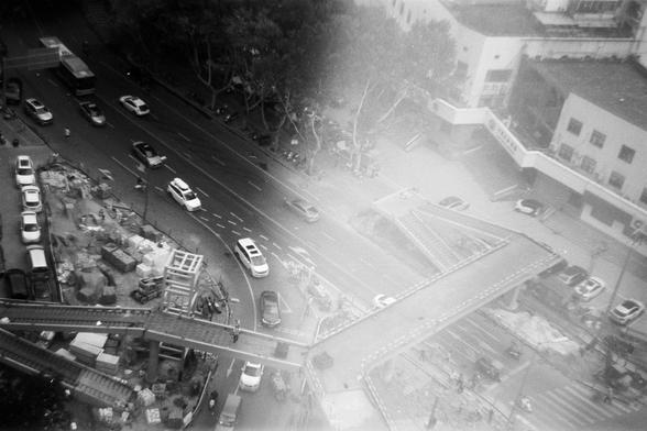 FUJIFILM NEOPAN F (FF)

English Alt Text: A black-and-white aerial image of a bustling city intersection. Cars, buses, and motorcycles move in various directions. A pedestrian overpass is under construction, with scaffolding and exposed beams. On the left side, construction materials and equipment are scattered. Buildings line the roads, and trees appear in the upper left. The photo captures the energy of urban life and infrastructure development.

中文替代文字：
这是一张黑白城市交叉路口的俯视图。汽车、公交车和摩托车在不同方向穿行。人行天…