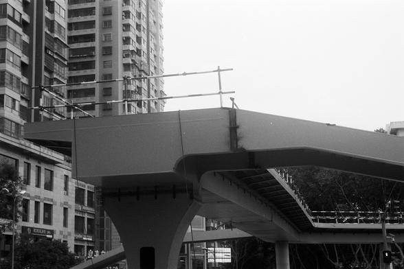 FUJIFILM NEOPAN F (FF)

English Alt Text: A black-and-white photo of an unfinished elevated pedestrian bridge in a city. The structure is made of metal and concrete, with railings and support columns visible. The bridge ends abruptly mid-air, not yet connected to another section. Surrounding the bridge are tall buildings and trees, blending urban development with nature. The image emphasizes architectural lines and contrasts between materials, capturing a moment of construction in progress.

中文…