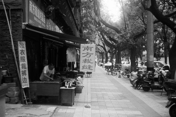FUJIFILM NEOPAN F (FF)

English Alt Text:
A black-and-white photo of a street in China. In the foreground, a tailor works at a sewing machine inside a small shop. Signs in Chinese characters hang outside, reading “Southern Tailor” and “Alter clothes and pant hems.” Trees, scooters, and pedestrians line the sidewalk. The scene captures everyday life and traditional craftsmanship in an urban setting.

中文替代文字：
这是一张黑白照片，拍摄于中国街头。前景是一家裁缝店，一位裁缝正在缝纫机前工作。店外挂着中文招牌，写着“南方裁缝”和“改衣服裤边”。人行道上有树木、电动车和行人。画面展现了城市环…