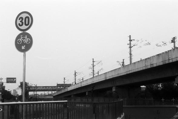 FUJIFILM NEOPAN F (FF)

English Alt Text:
A black-and-white photo showing an elevated railway track supported by concrete pillars. Overhead electric lines run above the track. On the left, two road signs are visible: one shows a speed limit of 30 km/h, and the other indicates a bicycle path below. Trees and a metal fence line the foreground. The image highlights urban transportation infrastructure and signage.

中文替代文字：
这是一张黑白照片，展示了城市交通基础设施。右侧是由混凝土柱支撑的高架铁路轨道，轨道上方有电线。左侧有两个交通标志：一个显示限速30公里/小时，另一个是自…