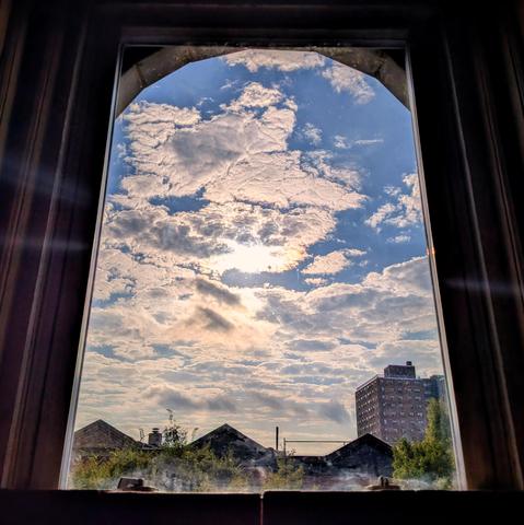 Looking through an arched window two hours after sunrise the sun is in the middle of the blue sky behind a dense pattern of white and grey clouds. Pointed roofs of Harlem brownstones are silhouetted across the street, and a taller apartment building can be seen in the distance. The green tops of two trees are on the bottom and right.