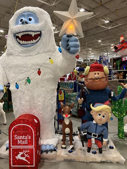 A store display of a festive holiday scene featuring large animated characters: a white yeti holding a star, a red-nosed reindeer, a boy dressed in blue with a tool, and a bearded man wearing a red hat.
