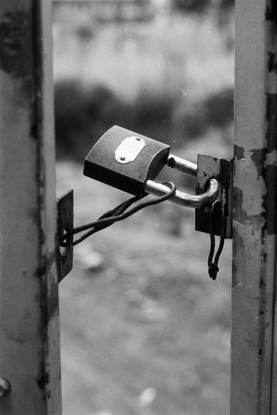 FUJIFILM NEOPAN F (FF)

English Alt Text: A close-up black-and-white image shows a weathered padlock securing two metal doors. The padlock is rusted and scratched, locked through thick metal loops. Two wires or cords are also threaded through the shackle, adding extra security or possibly serving another purpose. The background is blurred, suggesting an outdoor setting with faint outlines of buildings and vegetation. The image evokes a sense of age, security, and quiet tension, with the padlock…