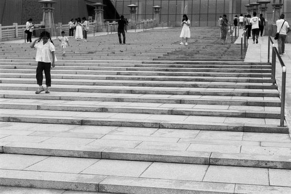 FUJIFILM NEOPAN F (FF)

English Alt Text: A black-and-white image shows a wide set of stone steps leading to a modern building with tall columns. Several people walk up and down the steps, some alone, others in groups. The building’s facade is smooth and contemporary, suggesting a public or cultural space like a museum. The handrails frame the steps, and the composition captures movement and structure. The monochrome palette adds a timeless, documentary feel, focusing on human activity in an ur…