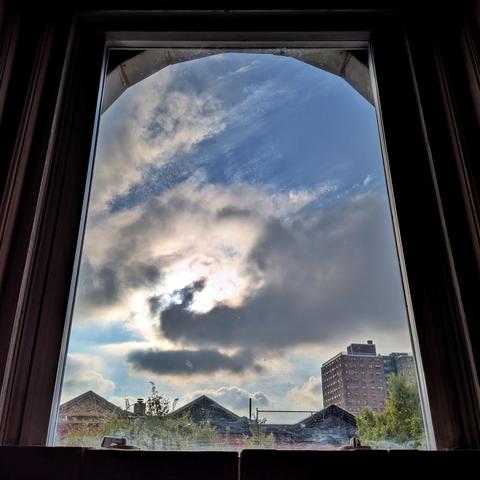 Looking through an arched window ninety minutes after sunrise the blue sky is filled with multiple layers of moving white and gray clouds, with large patches of blue showing through. Pointed roofs of Harlem brownstones are silhouetted across the street, and a taller apartment building can be seen in the distance. The green tops of two trees are on the bottom and right. The window is grimy on the bottom.
