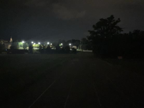 Looking down the straightaway of the running track, the middle school lit up in the distance. It’s nighttime with overcast skies and silhouetted trees.