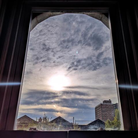 Looking through an arched window an hour and forty-two minutes after sunrise the blue sky is filled with complex layers of white, gray, and yellow clouds. The sun is above the houses, slightly to the left of center, and creating a lens flare as a curved horizontal white line across the frame. Pointed roofs of Harlem brownstones are silhouetted across the street, and a taller apartment building can be seen in the distance. The green tops of two trees are on the bottom and right. The window is gr…