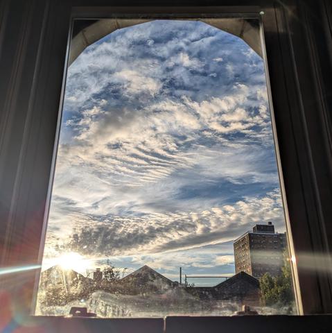 Looking through an arched window forty minutes after sunrise the boue sky is filled with complex layers of white and gray clouds. The sun is just above the houses on the left,  creating a lens flare as a curved horizontal white line across the frame. Pointed roofs of Harlem brownstones are silhouetted across the street, and a taller apartment building can be seen in the distance. The green tops of two trees are on the bottom and right. The window is grimy on the bottom.