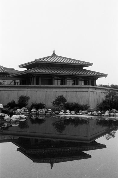 FUJIFILM NEOPAN F (FF)

English Alt Text:
A traditional East Asian-style building with a tiered, pagoda-like roof stands beside a calm body of water. The structure has slanted walls and overhanging eaves. In front, rocks and shrubs are arranged in a landscaped area. The water reflects the building, creating a symmetrical and serene composition. The architecture suggests Japanese or Chinese influence.

中文替代文字：
一座具有东亚风格的传统建筑坐落在宁静的水边，屋顶呈多层塔状，檐角上翘。建筑前方是布置有岩石和灌木的园林区域。水面清澈，倒映出建筑的轮廓，形成对称而宁静的画面。建筑风格可能受…