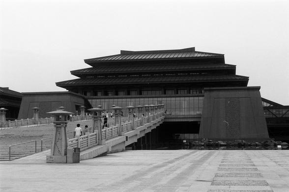 FUJIFILM NEOPAN F (FF)

English Alt Text:
A large building with East Asian architectural features, including a multi-tiered roof with upturned eaves, stands elevated behind a wide stone bridge. The bridge has decorative railings and lamp posts. A few people walk toward the entrance. The facade combines traditional stone carvings with modern glass panels. The image is in black and white, giving it a timeless, documentary feel.

中文替代文字：
一座大型建筑融合了东亚建筑元素，屋顶多层且檐角上翘。建筑高于地面，通过一座宽阔的石桥连接，桥上有装饰性栏杆和灯柱。几人正…