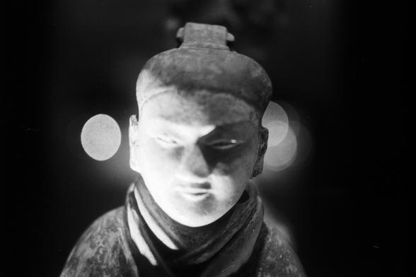 FUJIFILM NEOPAN F (FF)

English Alt Text:
A close-up of a stone or ceramic sculpture depicting a stylized human face. The figure wears a headpiece and a garment that wraps around the neck. The lighting is dramatic, casting shadows that emphasize the contours of the face and upper torso. The background is dark with soft circular light spots, creating a bokeh effect. The craftsmanship highlights cultural or historical significance, with attention to facial expression and texture.

中文替代文字：
这是一张石雕或…
