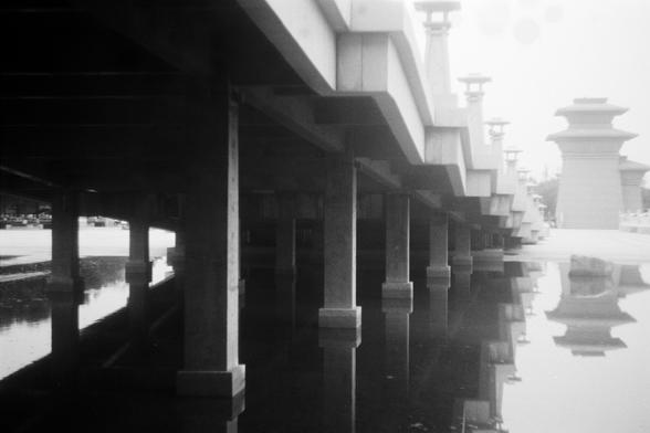 FUJIFILM NEOPAN F (FF)

English Alt Text: A black-and-white photograph taken from beneath a long bridge. The bridge stretches into the distance, supported by evenly spaced vertical columns that descend into a calm body of water. The water is so still that it mirrors the columns and underside of the bridge perfectly, creating a symmetrical visual effect. Along the top edge of the bridge are traditional lantern-style lamp posts, possibly East Asian in design, adding a cultural touch to the otherw…
