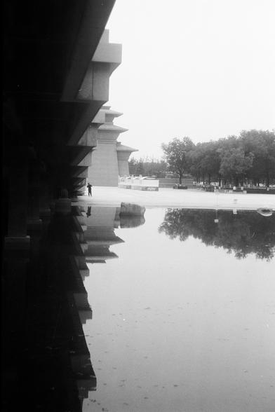 FUJIFILM NEOPAN F (FF)

English Alt Text: A black-and-white image of a modern architectural structure beside a large reflective pool. The building features angular overhangs and vertical columns, casting dramatic shadows. The water below reflects the entire structure, doubling its visual impact. In the background, trees and another uniquely designed building—possibly a museum or cultural center—add context. A lone person walks near the water’s edge, providing scale and a human element. The comp…