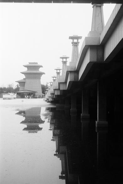 FUJIFILM NEOPAN F (FF)

English Alt Text: A black-and-white photo showing a modern bridge with stylized support columns extending into the distance. The bridge spans a body of water that reflects the structure and sky, creating a symmetrical scene. In the background stands a large, tiered tower resembling traditional East Asian pagodas, adding cultural depth. The image uses strong perspective lines and mirrored reflections to emphasize geometry and balance. The contrast between the sleek bridge…
