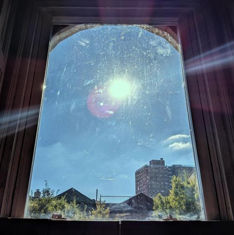 Looking through an arched window two-and-a-half hours after sunrise the sun is high in the blue sky, shining directly into the lens, creating a complex lens flare and illuminating the dirt and scratches on the glass.  There are a few small puffy white clouds in the lower right sky above the horizon. Pointed roofs of Harlem brownstones are silhouetted across the street, and a taller apartment building can be seen in the distance. The green tops of two trees are on the bottom and right. The windo…