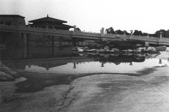 Kodak Double-X 5222 (FF)

English Alt Text:
A black-and-white photo of a serene scene featuring a traditional East Asian bridge and building. The bridge has ornate railings and decorative lamp posts. The building behind it has a tiered roof with upturned eaves, typical of classical Chinese or Japanese architecture. Below, a body of water reflects the structures above. Rocks and a paved area are visible in the foreground. The composition evokes a sense of peace and timelessness, possibly part of…