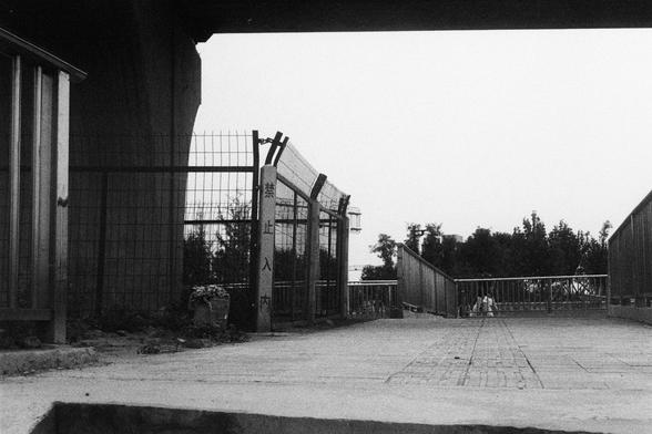 Kodak Double-X 5222 (FF)

English Alt Text:
A monochrome image of an outdoor pedestrian walkway, flanked by metal railings and fencing. A gate-like structure with vertical bars and mesh fencing stands in the center, possibly marking an entrance or exit. Behind the gate, trees and greenery suggest proximity to a park. Two small figures sit or crouch near the railing in the background. Overhead, a large structure—likely a bridge or overpass—casts shadows across the walkway. The concrete ground is…