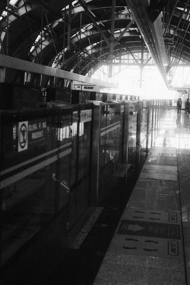 Kodak Double-X 5222 (FF)

English Alt Text:
A black-and-white photo of a sleek, modern train station platform. The architecture features a curved roof supported by exposed beams and large windows that allow natural light to flood the space. Glass barriers line the edge of the platform, separating passengers from the train tracks. Directional signs and floor markings indicate boarding zones and safety areas. A few people walk in the distance, adding a sense of quiet movement. The overall mood is…