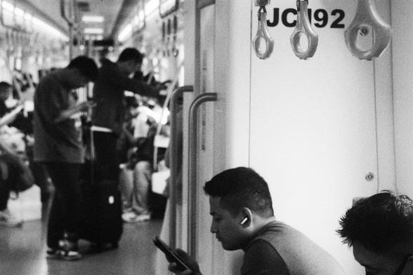Kodak Double-X 5222 (FF)

English Alt Text:
Inside a subway car, several passengers stand and sit in a well-lit, modern carriage. Most are absorbed in their smartphones, reflecting typical urban commuter behavior. Overhead hand grips hang from the ceiling, and one wall is marked with “JC192.” In the foreground, a seated man wears wireless earbuds and looks at his phone. Another seated person is partially visible. Standing passengers hold onto grips or lean against walls. The scene captures a qu…