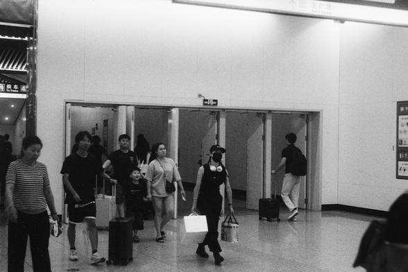 Kodak Double-X 5222 (FF)

English Alt Text:
In a public transportation terminal, travelers walk through a hallway with sliding glass doors. Some pull luggage, others carry shopping bags. A person in the foreground wears a face mask and cap, holding a drink and bags. Chinese signage above the doors suggests a location in a Chinese-speaking region. The image captures motion, routine, and the rhythm of travel.

中文替代文本：
在一个公共交通枢纽内，旅客穿过带有玻璃滑门的走廊。有些人拉着行李，有些人提着购物袋。前景中一人戴着口罩和帽子，手持饮料和购物袋。门上方的中文标识表明这是一个中…