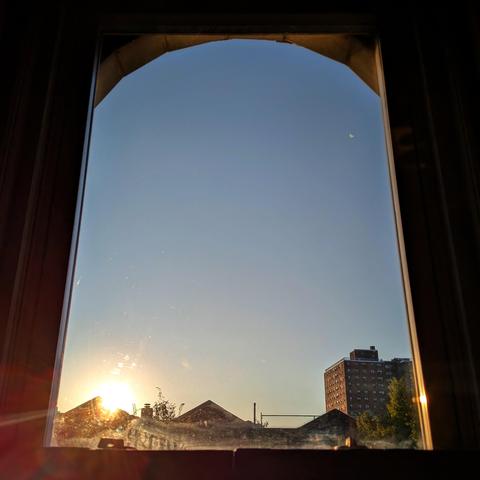 Looking through an arched window forty minutes after sunrise the sun is above the buildings on the left, making a lens flare. The sky is light blue and cloudless. Pointed roofs of Harlem brownstones are silhouetted across the street, and a taller apartment building can be seen in the distance. The window is grimy on the bottom.