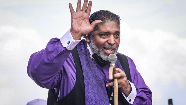 A man in purple liturgical garb with his hand up and his other hand holding a microphone and cane.
