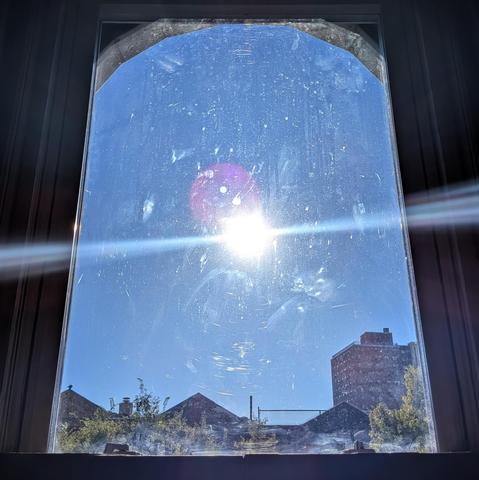 Looking through an arched window two hours after sunrise the sun is in the middle of the cloudless blue sky, shining directly into the lens, creating a complex lens flare and illuminating the dirt and scratches on the glass. Pointed roofs of Harlem brownstones are silhouetted across the street, and a taller apartment building can be seen in the distance. The green tops of two trees are on the bottom and right. The window is grimy.