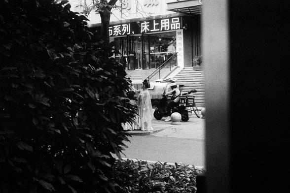 Kodak Double-X 5222 (FF)

English Alt Text:
A black-and-white photo taken through dense bushes. A person in light clothing stands near a motorbike with a delivery box on the back. The motorbike is parked on a sidewalk in front of a building with a staircase and glass entrance. Above the entrance, signage in Chinese reads “series, bedding products.” The hidden vantage point adds a candid, almost voyeuristic feel to the urban moment.

中文替代文字：
一张黑白照片，从浓密灌木中拍摄。一位穿浅色衣服的人站在一辆带配送箱的摩托车旁。摩托车停在人行道上，前方是一栋…